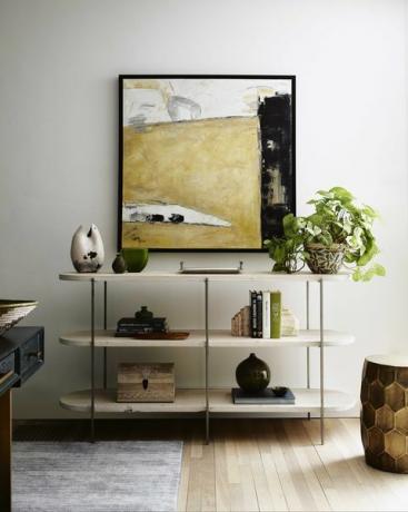 witte boekenplank, stapel boeken, gouden poef, kunst aan de muur, potplant