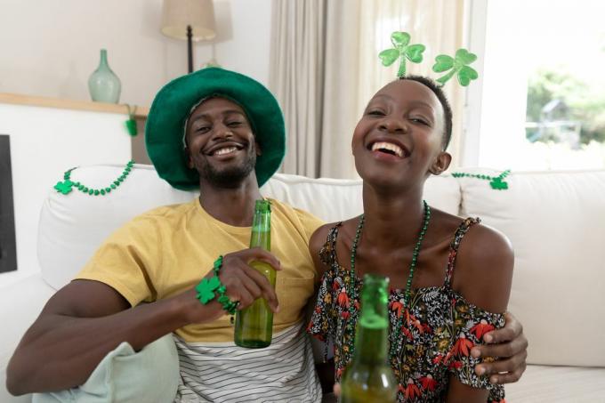 portret van lachend afrikaans amerikaans stel in st patricks dagkostuums met flessen bier geïsoleerd thuis blijvend tijdens quarantainevergrendeling