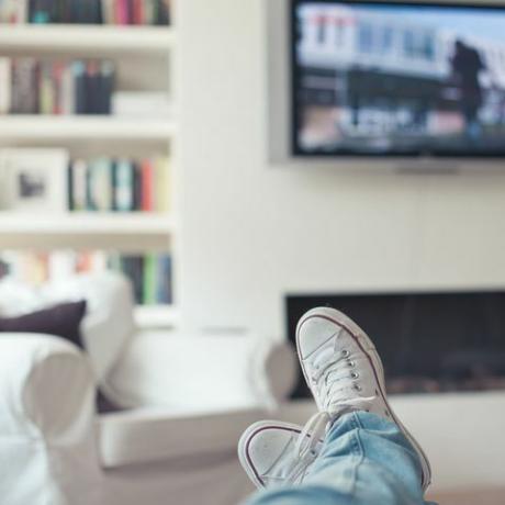 Foto van jeans en sneakers, voeten op de bank, met televisie, open haard, bank en boekenkast op de achtergrond.